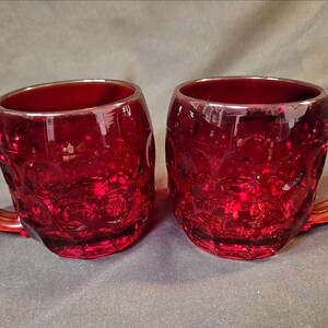May include: Two ruby red glass mugs with textured floral patterns. Each mug has a rounded body, a sturdy handle, and a wide opening. The mugs are made of a transparent glass that allows light to pass through, creating a vibrant appearance.
