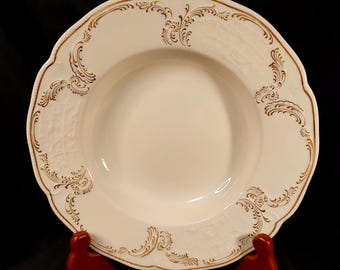 May include: A white, scalloped-edge soup bowl with a gold floral design around the rim. The bowl is set on a red stand against a black background. The bowl has a deep center and a decorative pattern.