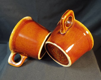 May include: Two brown ceramic mugs with a handle on each side. The mug on the right has the text "Hall's  Crestware  Made in  England" on the bottom.