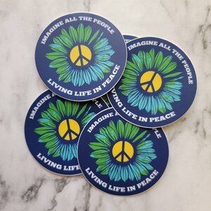 May include: A set of five round stickers with a blue background, a green and blue flower with a yellow peace sign in the center, and the text "Imagine All The People Living Life In Peace".