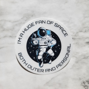 May include: A white round sticker with a black circle in the center. The black circle has a cartoon astronaut in a white spacesuit with a blue helmet giving a thumbs up. The astronaut is floating in space with stars. The text around the black circle reads "I'm a huge fan of space both outer and personal."