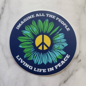 May include: A blue and white round sticker with a peace sign in the center. The peace sign is yellow and surrounded by a green and blue flower with a world map pattern. The text around the sticker reads "Imagine All The People Living Life In Peace."