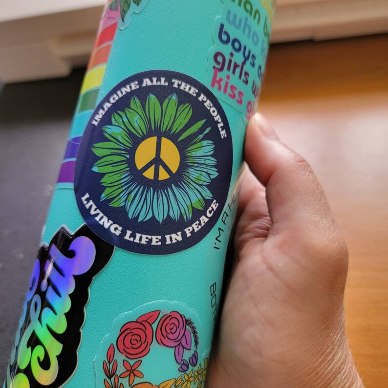 May include: A turquoise reusable water bottle with various stickers. One sticker features a peace sign inside a sunflower with the text "Imagine All The People Living Life In Peace".
