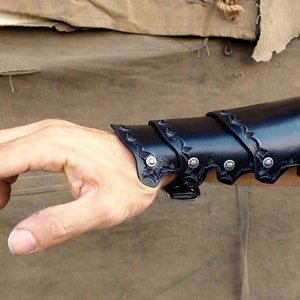 A leather bracer, shown being worn, set against a backdrop of worn canvas and corregated metal.