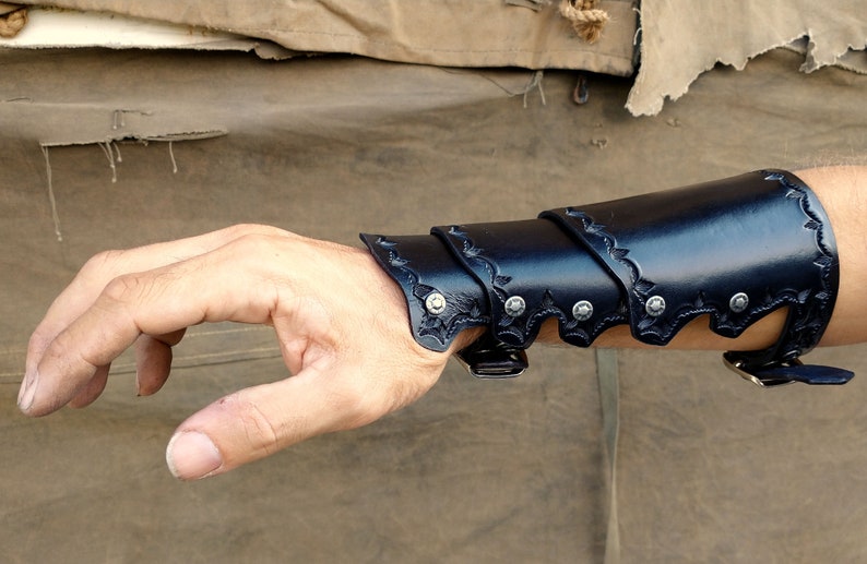 A leather bracer, shown being worn, set against a backdrop of worn canvas and corregated metal.