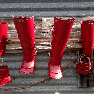 A set of bright red leather gauntlets, bracers, and leg greaves with added footplates. They are arranged against a metal garage with old barn wood rafters being used as a hanging rack.