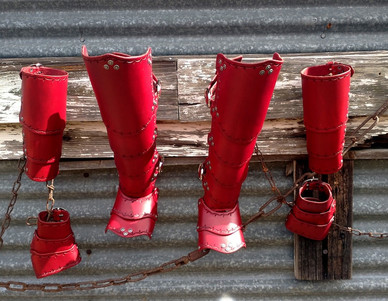 A set of bright red leather gauntlets, bracers, and leg greaves with added footplates. They are arranged against a metal garage with old barn wood rafters being used as a hanging rack.