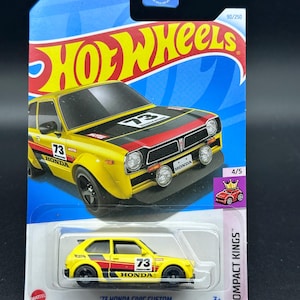 May include: A yellow 1973 Honda Civic Custom Hot Wheels die-cast car with black racing stripes, red and white accents, and the number 73 on the side. The car is on a blue and white background with the words "Hot Wheels" and "Compact Kings" printed on the packaging.
