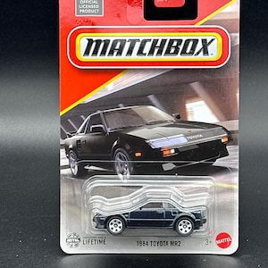 May include: A Matchbox die-cast toy car of a 1984 Toyota MR2, in a dark blue color, is displayed on a red and white card. The packaging includes the Toyota logo and the Matchbox logo. The car is approximately 3 inches long.