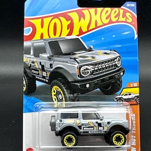 May include: A gray 2021 Ford Bronco Hot Wheels toy car with yellow wheels and black accents. The car is on a blue and white card with the Hot Wheels logo and the words "'21 Ford Bronco" and "HW Hot Wheels".