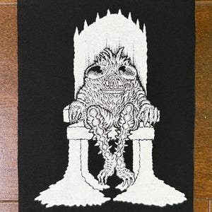 Könnte beinhalten: Schwarzer Leinwandkunstdruck mit einer weißen Illustration einer Kreatur auf einem Thron. Die Kreatur hat einen großen Kopf, spitze Zähne und stachelige Gliedmaßen. Der Thron hat eine hohe Rückenlehne mit spitzen Details und wird von zwei Beinen getragen.