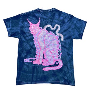 以下が含まれることがあります： ネイビーブルーのタイダイTシャツ。ピンクと紫色の猫のイラストが描かれています。猫は詳細なデザインで直立しています。デザインには、猫の体を包む一連の線が含まれています。