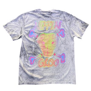 Puede incluir: Camiseta gris de manga corta con un patrón texturizado. La parte posterior de la camiseta presenta un diseño de calavera y huesos cruzados de neón con las palabras "LOFTIN GLOO" en una fuente colorida de estilo grafiti.