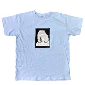 May include: A light blue short-sleeve t-shirt featuring a rectangular black patch on the chest. The patch contains a white line-art illustration of a spiky, creature-like figure standing on a rocky surface, rendered in a detailed, sketch-like style.