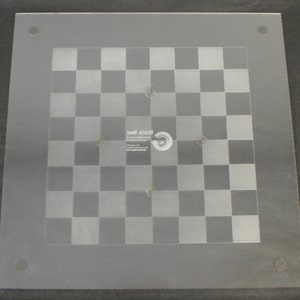 May include: A clear glass chessboard with a frosted checkerboard pattern. The board has eight small, round, silver-colored dots around the perimeter and a larger dot in the center. The text "Magic Game" is printed in the center of the board.