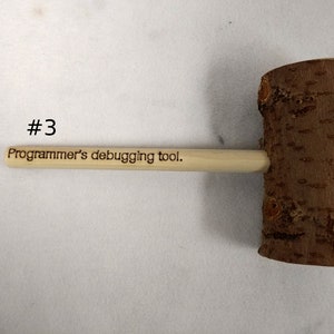 Pode incluir: Um martelo de madeira com uma al&ccedil;a que diz "Ferramenta de depura&ccedil;&atilde;o para programadores". O martelo &eacute; feito de madeira natural com uma textura de casca &aacute;spera.