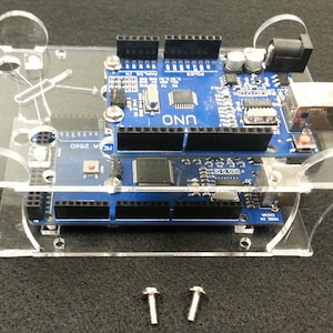 Könnte beinhalten: Ein transparentes Acryl-Gehäuse mit zwei Schrauben für eine Arduino Uno Mikrocontroller-Platine. Die Platine ist blau und hat den Text "UNO" aufgedruckt.