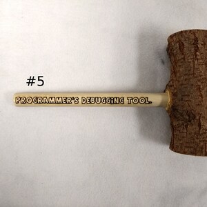 Pode incluir: Um martelo de madeira com acabamento em casca marrom natural. O cabo &eacute; feito de madeira clara e tem o texto "PROGRAMMER'S DEBUGGING TOOL." gravado.