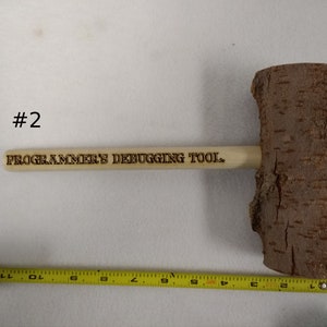 Pode incluir: Um martelo de madeira com acabamento em casca natural e uma al&ccedil;a que diz "PROGRAMMER'S DEBUGGING TOOL."