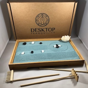May include: A wooden desktop zen garden with a blue sand base, black and white stones, a rake, and a small white flower. The box is labeled "Desktop Tranquility".