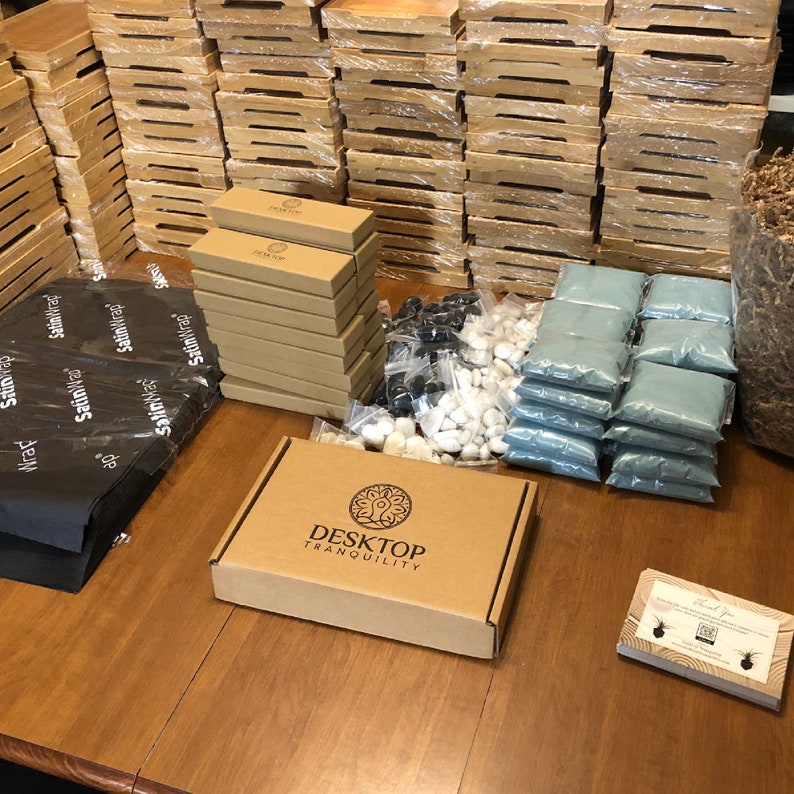 May include: A brown cardboard box with the text "Desktop Tranquility" printed on the top. The box is surrounded by various items, including small bags of sand, white and black stones, and a stack of brown cardboard boxes.