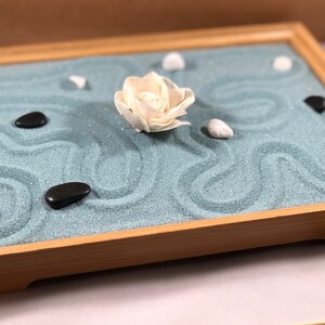 May include: A wooden tray with a blue sand garden. The sand is sculpted into waves and there are white and black stones and a white flower on top.