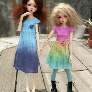 May include: Two dolls wearing colorful ombre dresses. The doll on the left has red hair and is wearing a blue dress with a purple flower. The doll on the right has blonde hair and is wearing a pink and yellow dress with a pink flower.