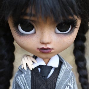 Könnte beinhalten: Nahaufnahme einer Puppe mit schwarzen Zöpfen. Die Puppe hat dunkle Augen, lange Wimpern und einen ernsten Gesichtsausdruck. Sie trägt einen grauen Blazer, einen schwarzen Pullover, ein weißes Hemd und eine schwarze Krawatte. Der Text "WEDNESDAY BELLADOLLADOLLS" ist sichtbar.