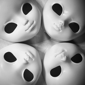 May include: Four blank white doll heads with black painted eyes and mouths. The doll heads are arranged in a circle on a white surface.