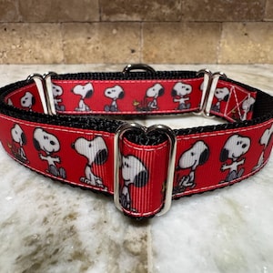 Könnte beinhalten: Ein rotes Hundehalsband mit einem sich wiederholenden Muster von Snoopy-Figuren. Das Halsband hat eine schwarze Rückseite und silberfarbene Metallbeschläge. Das Snoopy-Design ist in Weiß und Schwarz.
