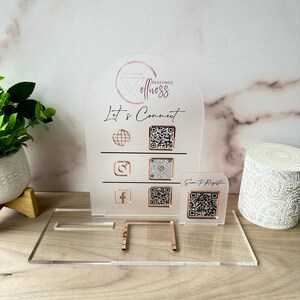 May include: Clear acrylic sign with rose gold accents. The sign features the text "Redefined Wellness" and "Let's Connect" with QR codes for social media and registration. A small stand supports the sign.
