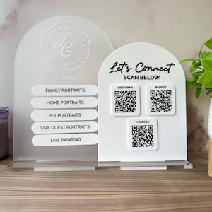 Op de afbeelding: Een wit acryl bord met de tekst "Let's Connect" en QR-codes voor Instagram, Website en Facebook. Het bord vermeldt ook portretservices: Familie, Thuis, Huisdier, Live Gasten en Live Schilderij. Het bord staat op een houten oppervlak.
