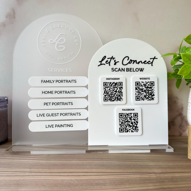 Op de afbeelding: Een wit acryl bord met de tekst "Let's Connect" en QR-codes voor Instagram, Website en Facebook. Het bord vermeldt ook portretservices: Familie, Thuis, Huisdier, Live Gasten en Live Schilderij. Het bord staat op een houten oppervlak.