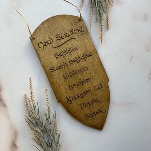 May include: Wooden sign with the words "Now Serving" and a list of meal times, including "Breakfast", "Lunch", and "Dinner". The sign is a light brown color and has a decorative shape. A gold string is attached for hanging.