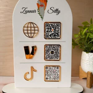 May include: A white arched acrylic sign featuring the text Zunnar Silty and a colorful illustration of a person carrying a basket. It includes three gold-colored social media icons and three corresponding square QR codes on a stand.