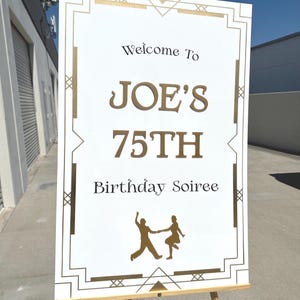 May include: A white sign with gold Art Deco style border and text. The sign reads "Welcome To JOE'S 75TH Birthday Soiree" in gold and black lettering. A gold silhouette of a dancing couple is at the bottom.