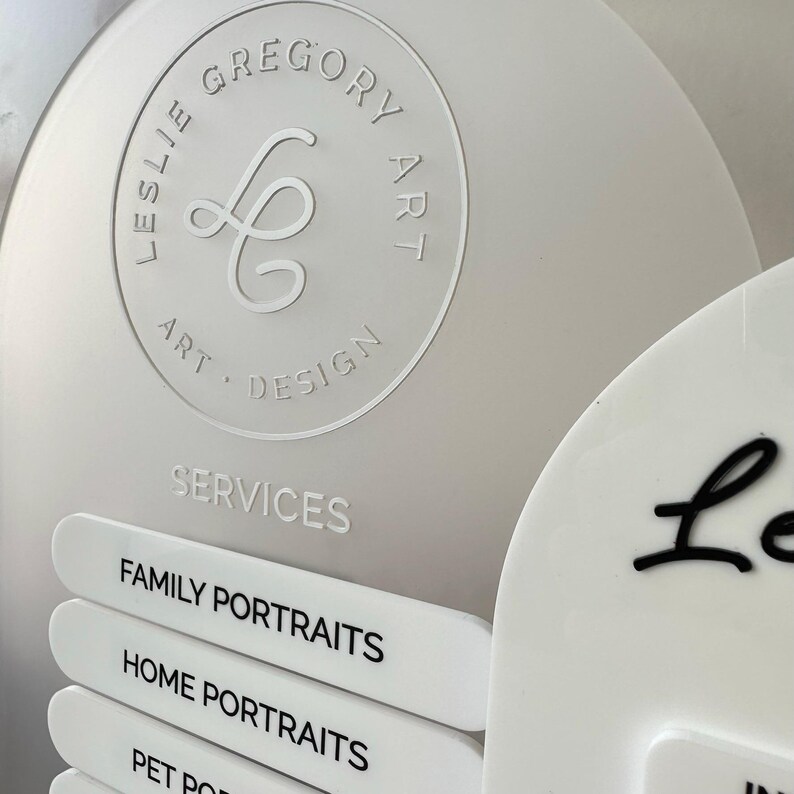 Op de afbeelding: Close-up van een lichtgrijs bord met de tekst "LESLIE GREGORY ART DESIGN" in een cirkelvormig ontwerp. Hieronder is het woord "SERVICES" zichtbaar, met labels voor "FAMILY PORTRAITS", "HOME PORTRAITS" en "PET PORTRAITS".