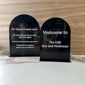 May include: Two black acrylic signs with rounded tops. The sign on the left says "By Appointment only! To book please text or call: 902-214-0932 email: theeditspaandwellness@gmail.com Treatment in session please do not disturb." The sign on the right says "Welcome to The Edit Spa and Wellness!"
