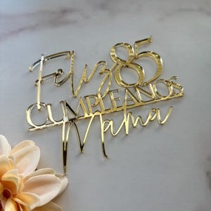 May include: A gold-colored cake topper with the words "Feliz Cumpleaños Mama" and the number "85". The topper is made of a shiny, reflective material and is designed to be placed on top of a cake. The text is in a script font.