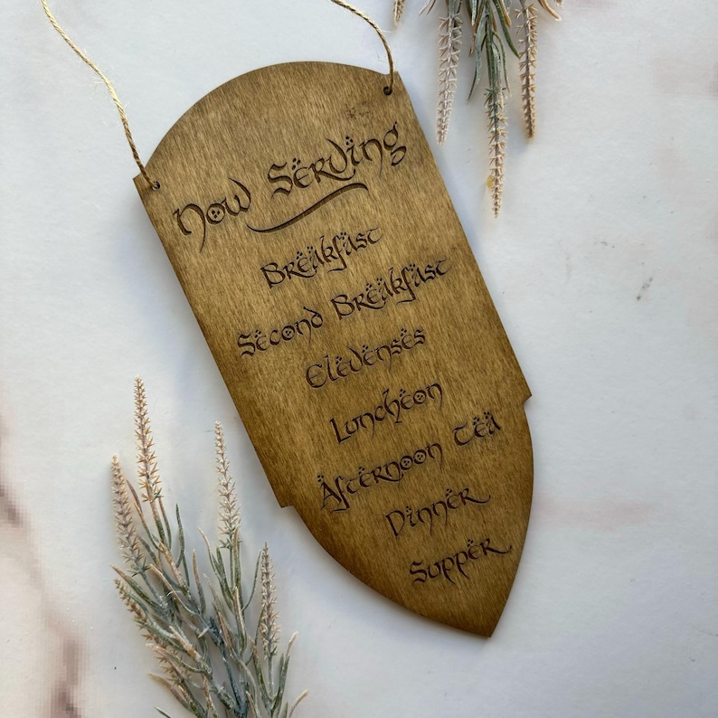 May include: Wooden sign with the words "Now Serving" and a list of meal times, including "Breakfast", "Lunch", and "Dinner". The sign is a light brown color and has a decorative shape with a string for hanging.