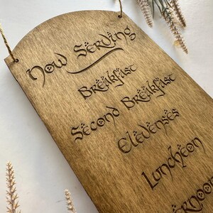 May include: Wooden sign with the words "Now Serving" and meal times etched into the wood. The sign is rectangular with a rounded top and a hole for hanging. The text is in a decorative font.