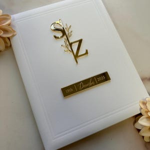 May include: A white invitation card with a gold-colored monogram of the letters "SZ" and a sprig of leaves. A gold rectangular plate displays the date "18th | December | 2025". The card has a raised border.