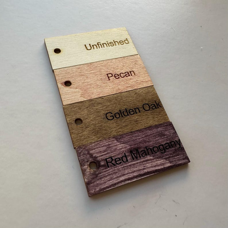 May include: A wooden color swatch card featuring four different wood stain colors: Unfinished, Pecan, Golden Oak, and Red Mahogany. Each swatch has a small hole and the color name engraved on it.