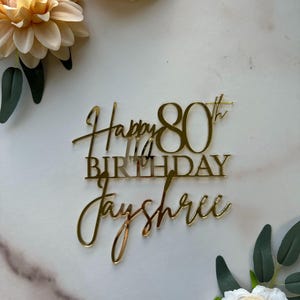 May include: Gold glitter cake topper with the text "Happy 80th Birthday Jayshree".