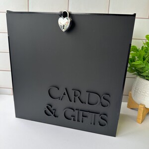 May include: A square black box featuring the text CARDS & GIFTS in raised black lettering on the front. The top of the box is secured with a silver-colored heart-shaped padlock. The box has a smooth, matte finish and a clean, modern design.