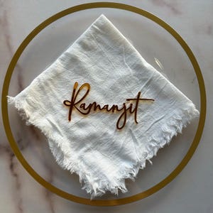 May include: A clear glass charger plate with a gold rim, holding a white linen napkin. A gold-colored acrylic name tag that reads "Ramanjit" is placed on the napkin. The napkin has a frayed edge.