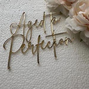 May include: A gold-colored cake topper with the words "Aged to Perfection" in a cursive font. The topper is placed on a white surface with a textured pattern. Soft pink flowers are in the background.