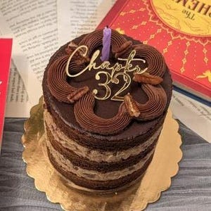 May include: A layered chocolate cake with chocolate frosting and pecan accents. A gold cake topper reads "Chapter 32" with a purple candle. The cake sits on a gold scalloped cake board.