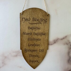 May include: Wooden sign with the words "Now Serving" and a list of meal times, including "Breakfast," "Second Breakfast," and "Supper." The sign is shield-shaped and hangs from a natural twine string.