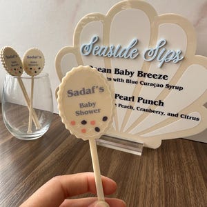May include: A light beige baby shower drink stirrer with the text "Sadaf's Baby Shower" and decorative dots. A clear glass and a seashell-shaped sign with "Seaside Sips" are also visible.